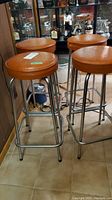 Four chrome-base bar stools with brown round cushioned seats arranged around a small bar cabinet with liquor bottles and glassware in the background.