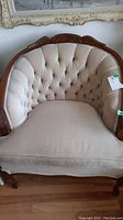Front view of beige tufted chair with carved wood frame showing tufted seat, backrest, and wood top rail and arms.
