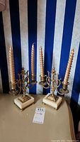 Pair of metal and stone 3-arm candle holders with 3 twisted gold candles each on a beige surface against a blue and white striped background.