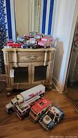 Photo showing a collection of vintage toy cars and trucks on a small cabinet and floor including trucks, fire trucks, and piggy banks.