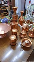 Photo showing the full collection of copper ware on a wooden table including bowl, vases, tea set components, and shakers.