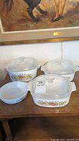 Four CorningWare casserole dishes with vegetable pattern and glass lids on a wooden table beneath a framed painting.