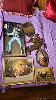 Six wall art items are laid out on a purple bedspread: Elvis Presley print, 1932 Dusenberg car print, knight metal wall art, oval mirror, oval woman art print, and moose wood art signed by H Nadeau.