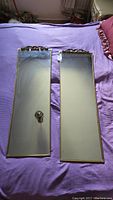 Two long rectangular mirrors with gold-toned thin frames and detailed carved decorative tops placed on a purple surface