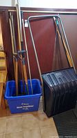 Overall view of the lot showing several wooden-handled garden shovels and rakes placed inside a blue plastic container, accompanied by a large black snow shovel with metal handle.