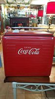 Front view of vintage Coca-Cola metal cooler showing red color and white logo, with upper wooden shelf on top.