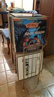 Collection of vinyl records stored vertically on a gold-toned metal stand with mid-century modern styling in a room with tiled floor and mid-century furniture.