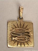 Front view of the gold fish pendant showing square shape, textured sunburst background and two raised fish figures.
