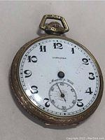 Full front view of antique pocket watch with white dial, black Arabic numerals, no hands, gold filled decorative case.