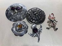 Overview of five Scottish brooches including thistle round brooches and a Highlander figure brooch on a white background.