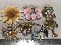 All seven vintage ladies brooches arranged on a surface with a ruler showing size reference, including floral designs, a cluster of pearls, a poodle, and a bow brooch.