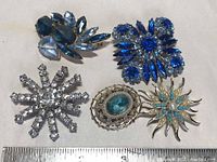 Overview of all five vintage rhinestone brooches arranged on a beige surface with a ruler for scale.