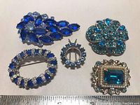 Brooches arranged on a white surface with a ruler for scale, showing five vintage brooches with blue rhinestones and different designs.