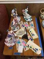 Wooden table with 4 porcelain Asian figurines, 8 fabric dolls laid out, and folding fan near center.