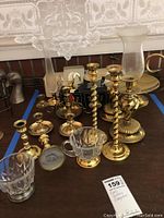 Photo showing grouped brass candle holders, cut glass pieces, and a glass globe on a dark surface with lace curtain background.
