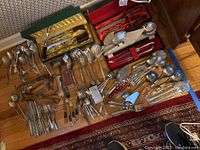 Wide view of the entire lot showing multiple silver plated and stainless steel serving utensils and flatware spread out on a wooden floor with different boxes and cases.