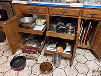 Kitchen open cabinet with various cookware including copper bottom Revere Ware pots with lids, glass Vision pot with lid, Orchard Royale casserole box, and metal container pieces placed inside and in front of cabinet. Several cutting boards seen to the side.