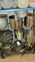 Photo of multiple large and medium pots and pans on a metal wire rack. Includes commercial style stock pots and pans, absence of some lids noted.