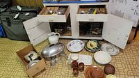Wide view showing boxed items, ceramic pitcher, roasting pan with lid, salt and pepper shakers, brown picnic plates, floral plates, and various bowls and cups on floor