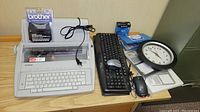 Brother GX-6750 electric typewriter with typewriter ribbon cartridge, computer keyboards, mousepad with calculator, wired mouse, battery-operated clock, and batteries displayed on a wooden desk