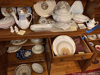 Wood cabinet shelf with assorted white china including teapot, plates, soup tureen with cracked handle, creamers, and salt/pepper shakers
