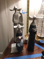 Three Lladro figurines with a tape measure showing heights from about 7 to 11 inches, displayed on a wooden surface next to a lamp.
