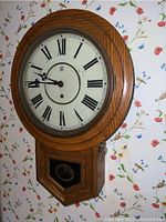 Full view of oak wood framed regulator wall clock showing the dial, hands and pendulum box on floral wallpaper background.