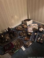 Overview of silver plated items laid out on carpet including tea set, trays, serving bowls, utensils, napkin rings and corn cob holders
