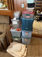 Stack of various plastic storage totes including large blue Rubbermaid tote, several clear totes with lids, and black basket, with used newsprint paper sheets stacked in front.