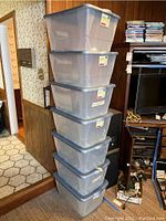 Stack of seven large gray Homz storage totes with lids, stacked vertically against a wood-paneled wall near a doorway and entertainment unit.