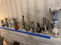Full shelf view of 16 assorted vases arranged side by side. Materials include pewter, porcelain, china, and glass in various colors and heights.