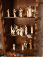 Three wooden shelves displaying 19 Willow Tree style figurines in various poses on middle and top shelves with angels, praying figures, and human forms in neutral tones.