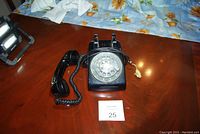 Black rotary dial phone placed on a wooden table with coiled handset and attached beige phone plug visible.