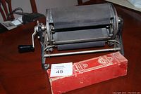 Side view of vintage No.30 Mimeograph machine with crank handle and accompanying red ink box on wooden table.