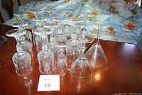 16 crystal glasses arranged on a wooden table with floral cloth backdrop. Includes various stemware types all clear crystal with cut or etched patterns.