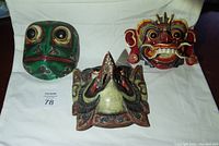Three wooden carved masks arranged on a white cloth: a green mask, red fiend mask, and bird beak mask.