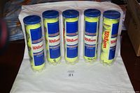 Five unopened packs of Wilson tennis balls arranged upright, packaged in clear plastic tubes with blue lids.