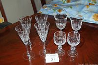 Photo showing all 8 crystal wine glasses arranged on a wooden table with a blue floral tablecloth in the background, showcasing the different glass shapes and cut patterns.