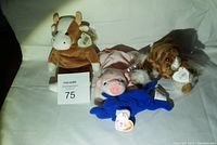 Four Beanie Babies plush animals arranged on a white sheet: a brown and white horse, a pink pig, a blue elephant, and a brown and white cow each with TY tag visible.