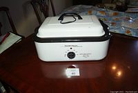 Hamilton Beach 18 quart roaster oven with black handle on white lid and black trim around the base, sitting on a wooden table.