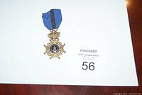 Full view of the medal with attached blue ribbon, showing the crown and star shape on a white background with auction paper.