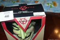 Front view of the boxed V for Vendetta deluxe mask, showing the mask's face and the packaging design with logos and text.