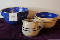 Three stoneware items: large blue mixing bowl, natural colored pitcher with blue bands, smaller blue glazed bowl with metal handle