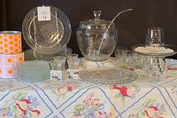 Full lot on floral cloth background including punch bowl with lid and ladle, cups, glass plates, cake plates, stemless wine glasses, bud vases, tins, and tablecloth.