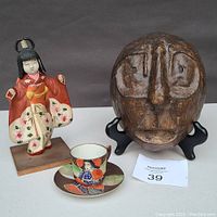 Photo showing the full lot with mask on black stand, Asian doll figurine in colorful kimono, and Japanese Moriage teacup with saucer.