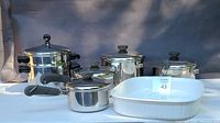 Overall view of the entire cookware set including white Corning Ware casserole and five stainless steel Revere Ware pots with lids and steamer insert.