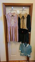 Two vintage ladies' dresses hanging on a door: pink silk sleeveless dress with lace front and black bow, and black silk dress with lace collar and velvet skirt. Baby clothes jacket visible below.