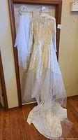 Full view of wedding gown hanging with attached long lace sleeves, lace overlay, and tulle train displayed alongside crown veil made of beads, pearls, crystals, and tulle layers.