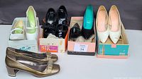 All 5 pairs of vintage women's high heels arranged in line, some with original boxes visible. Colors include white, black, and green/gold. Various styles including low and high heels with leather and patent leather materials.