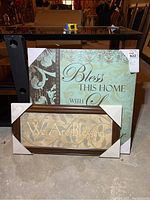 Two canvas prints placed upright against a black piece of furniture. The larger canvas is unframed with corner protectors and reads 'Bless This Home with Love & Laughter.' The smaller framed canvas reads 'We Are Blessed.'